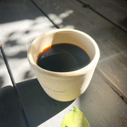 A beige NEW MIND ceramic intention cup filled with dark Chaga coffee, resting on a wooden table outdoors.