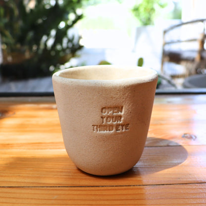 A beige hand-made ceramic NEW MIND intention cup engraved with 'Open Your Third Eye', on a wooden table.