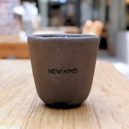 A hand-made ceramic intention cup in charcoal color with the engraved message 'NEW MIND' and 'Open Your Third Eye' displayed on a wooden surface.
