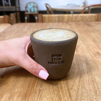 Hand holding a hand-made charcoal ceramic NEW MIND intention cup filled with Chaga latte, engraved with 'Open Your Third Eye'.
