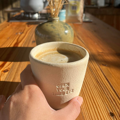 Hand holding a beige NEW MIND ceramic intention cup filled with Chaga coffee.