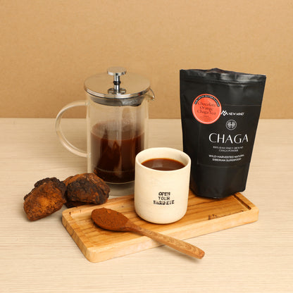 Chocolate Orange Chaga Tea served in a cup with a French press, wooden spoon, Chaga chunks, and the product bag on a wooden tray.