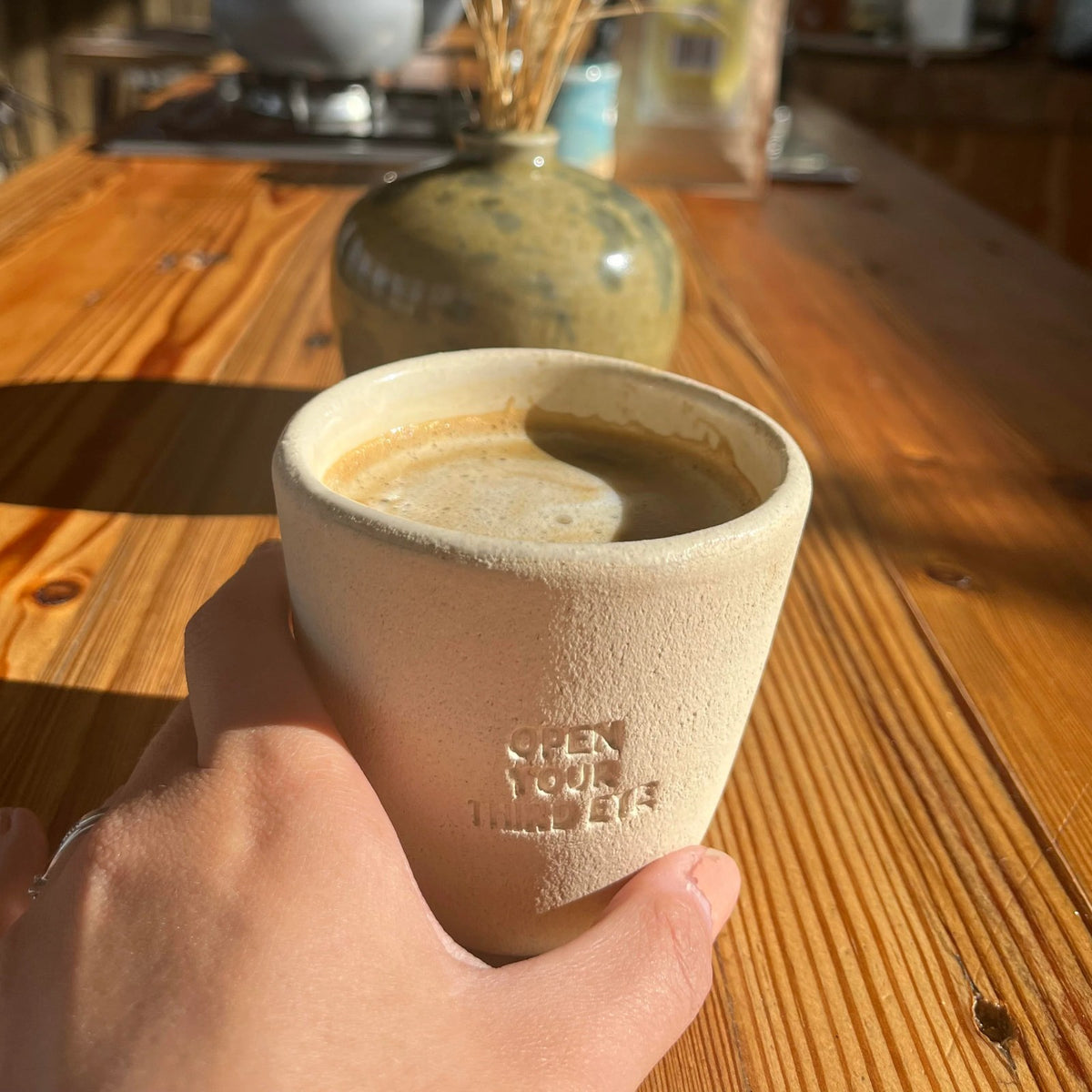 Hand holding the NEW MIND ceramic intention cup with a latte, Open Your Third Eye engraving facing out, on a sunny wooden table